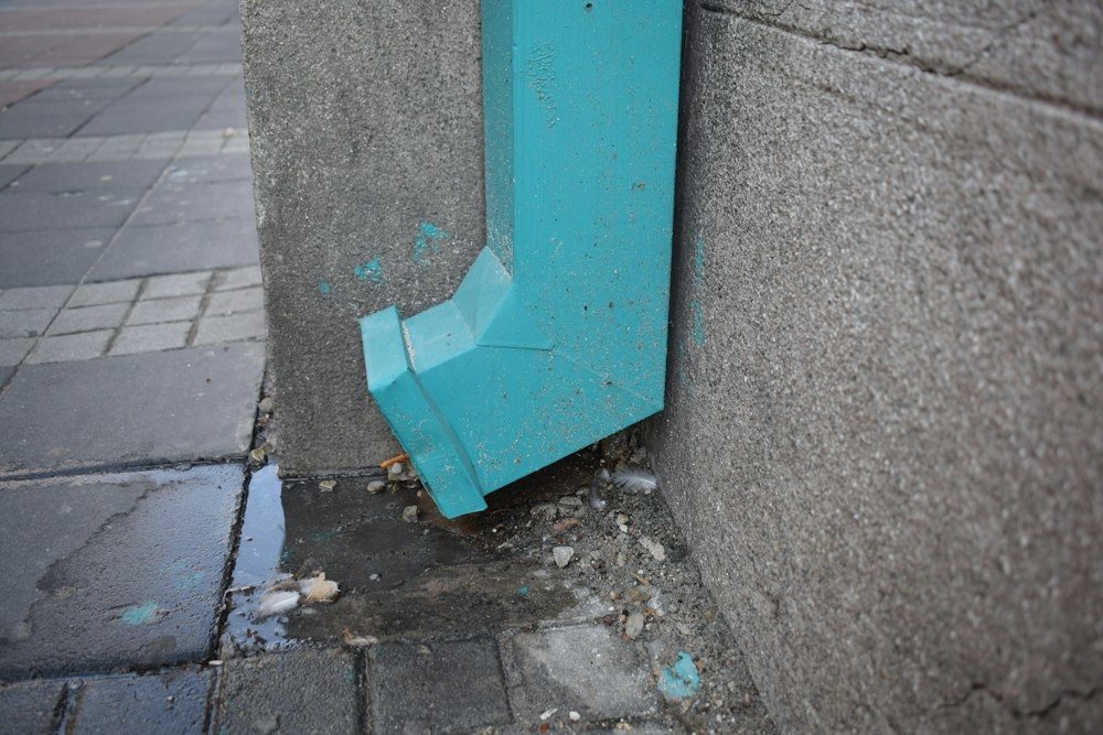 A downspout elbow at the base of a concrete wall with wet pavement below it